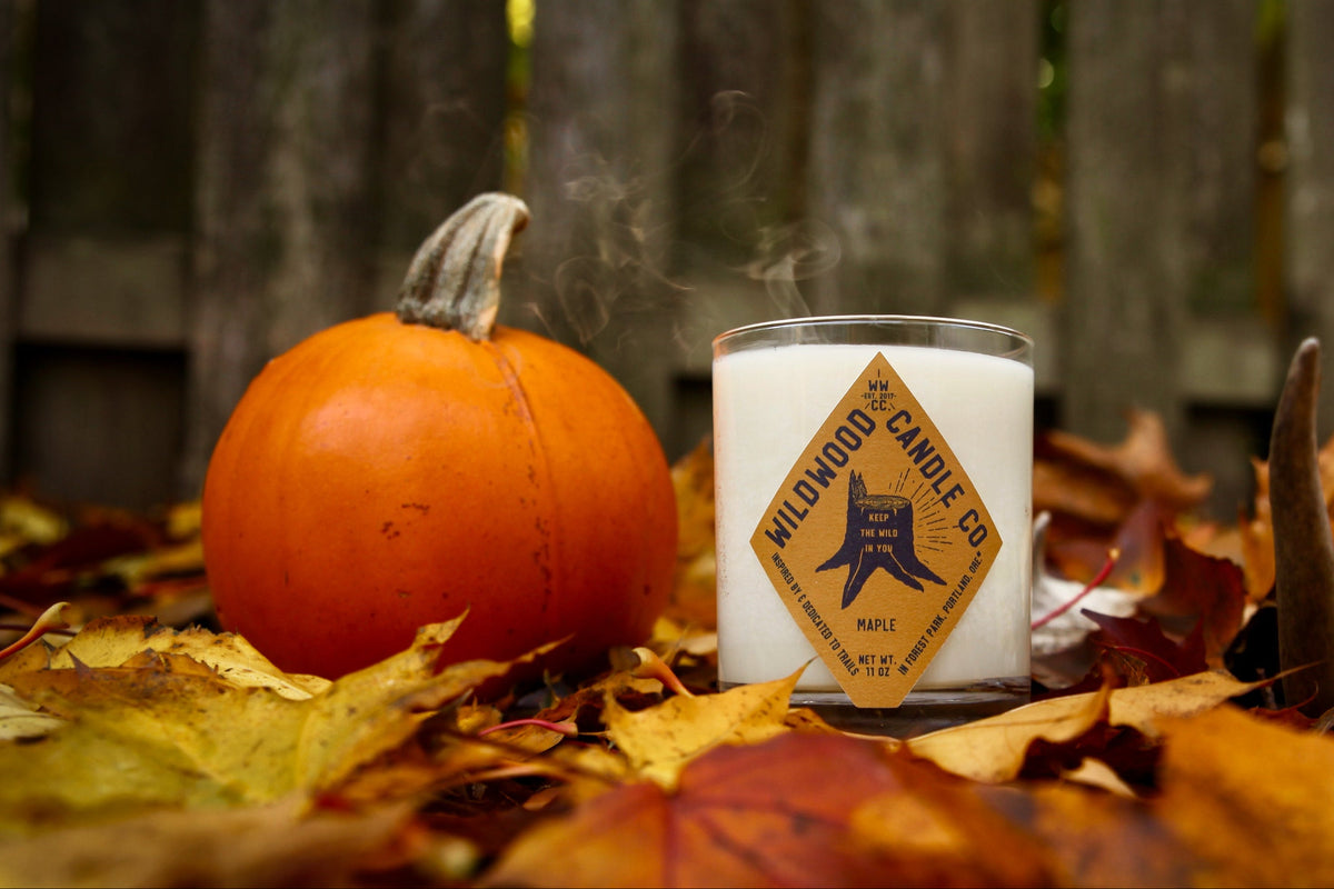 A white candle jar labeled 'Wildwood Candle Co.' sits outdoors on a bed of fallen autumn leaves next to a small orange pumpkin, with a wooden fence in the background.