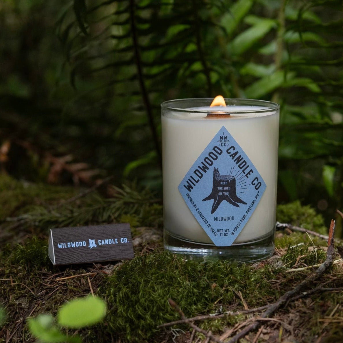Candle with a label on a forest floor with ferns in the background